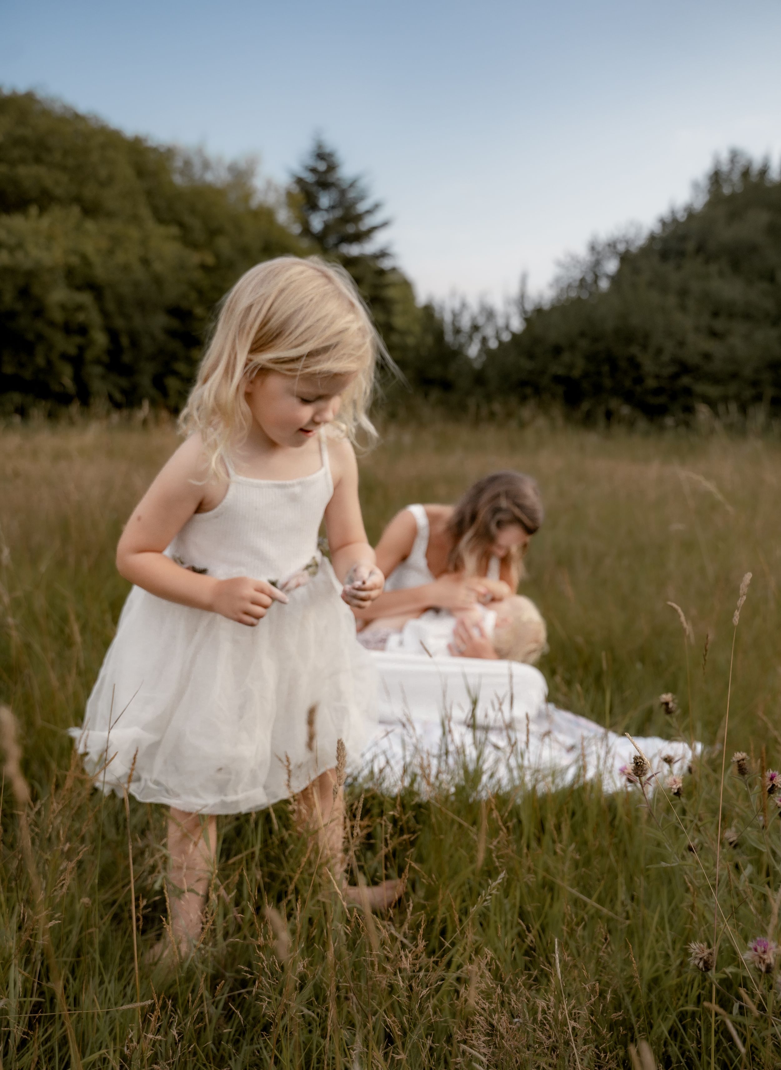 Two people in white dresses sit together in a serene meadow with tall grass during golden hour.