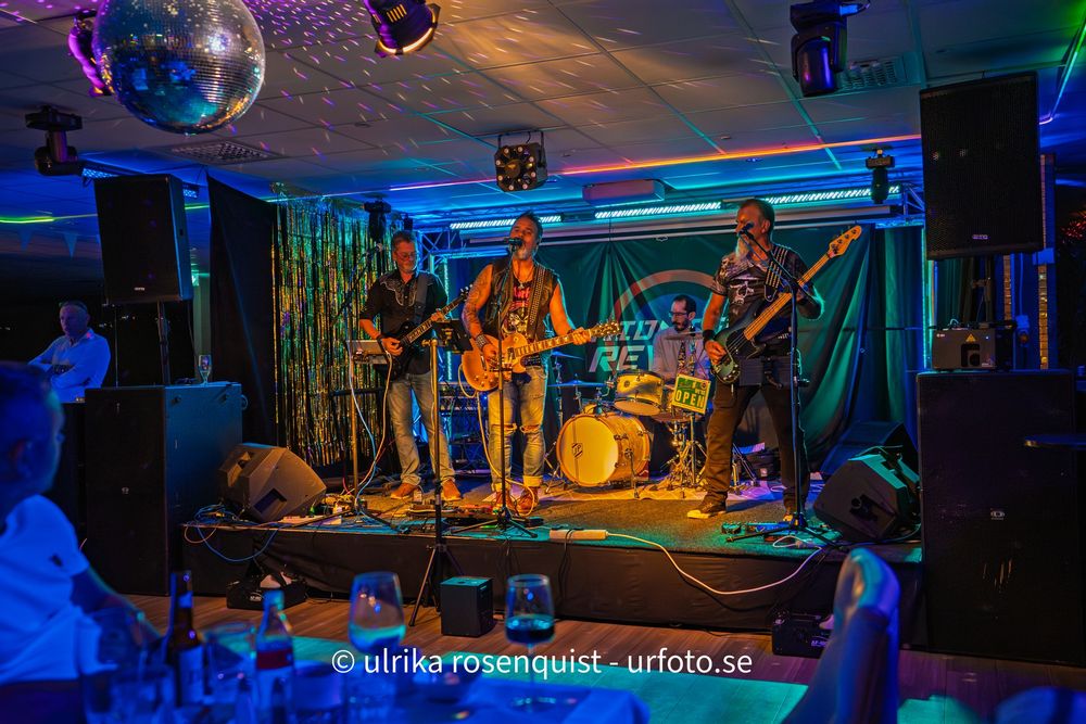 A live band performs on stage in a nightclub with blue lighting and a disco ball overhead.