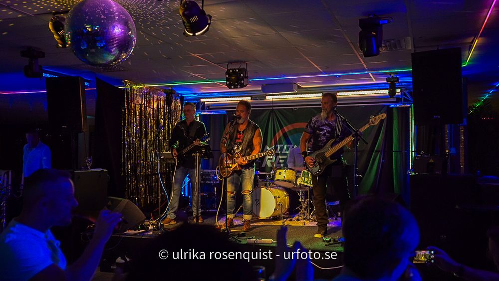 Musicians perform on stage in a dark venue with colorful purple and blue lighting effects.