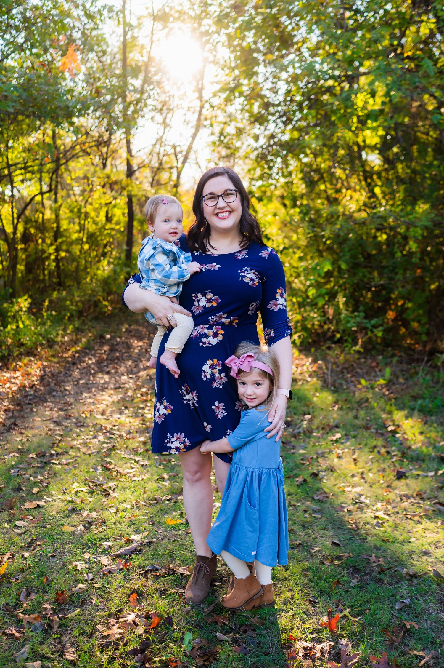 Mom holds infant while preschool daughter hugs mom's leg during outdoor family photos. family wearing blue with sun shining from behind.