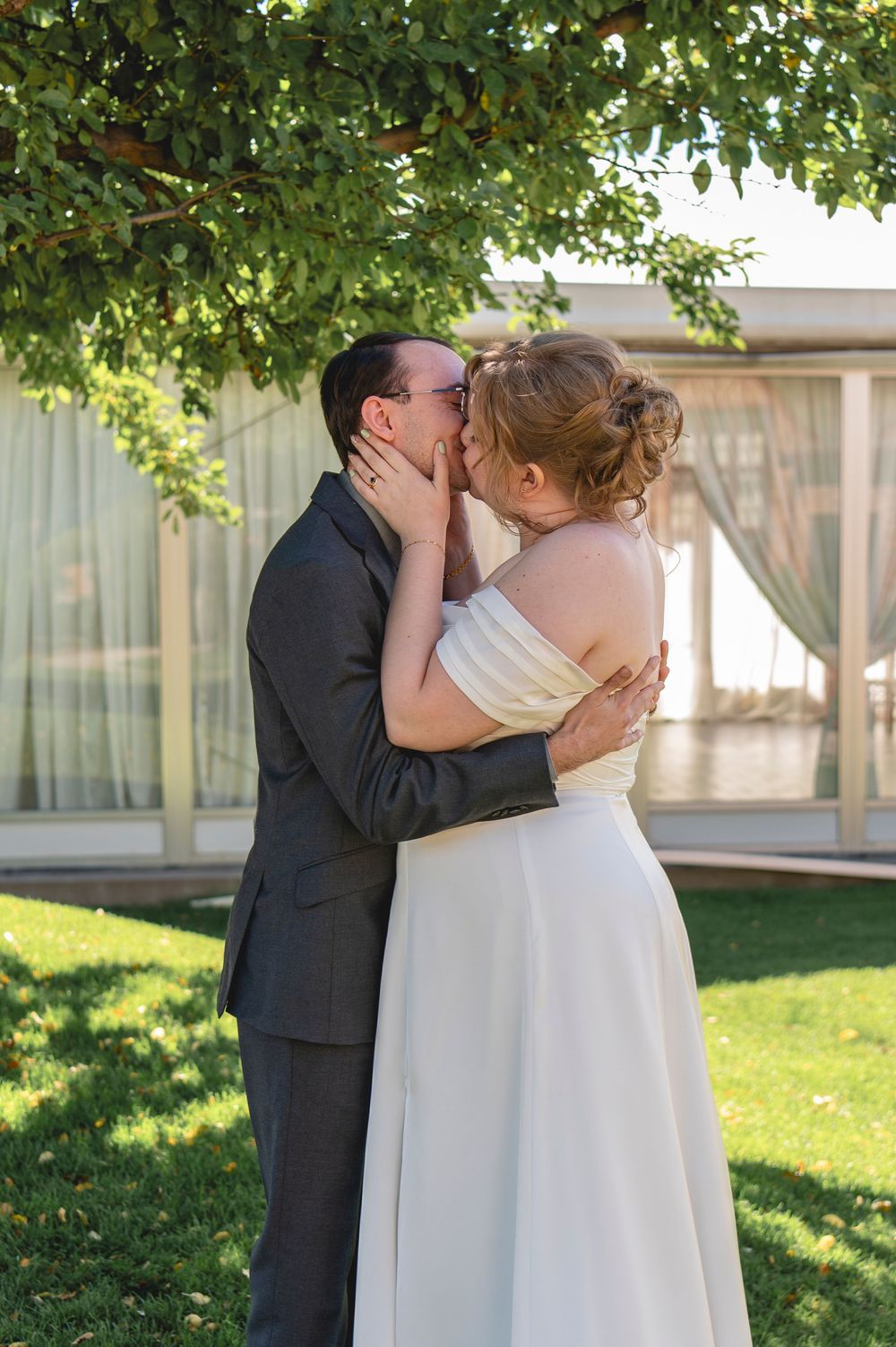 A couple embrace and kiss in a sunlit garden setting wearing traditional wedding attire.