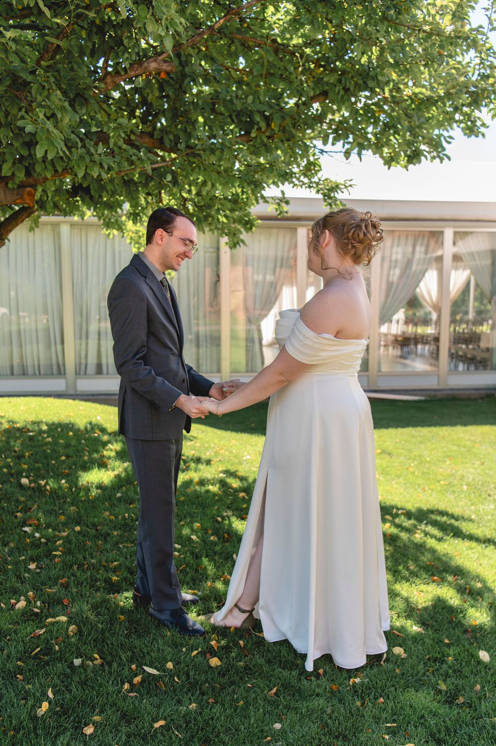 A romantic first look moment takes place outdoors with green trees and scattered leaves in the background.