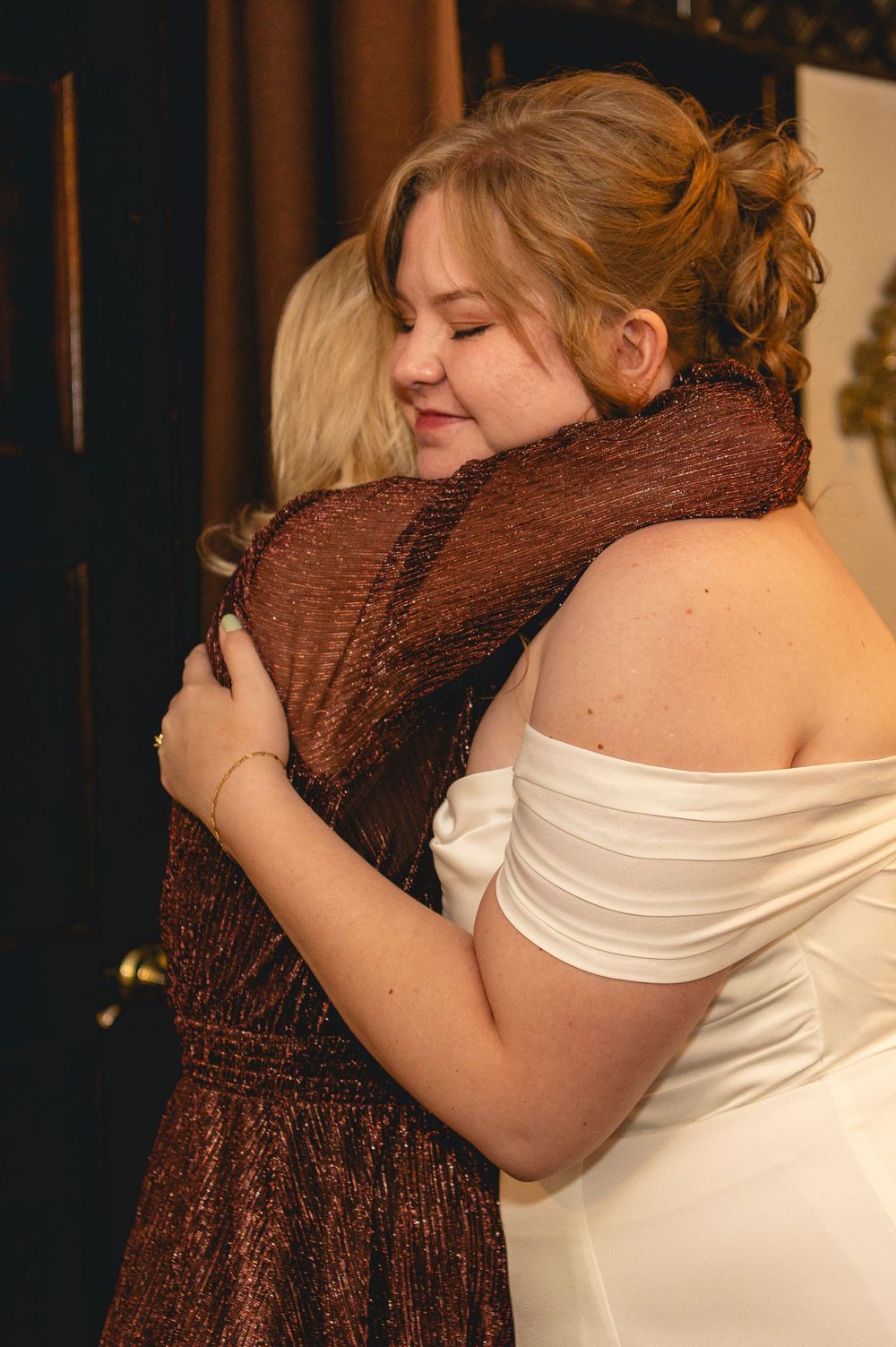 Two people share an emotional embrace at what appears to be a formal event, one wearing a white dress and one in brown.