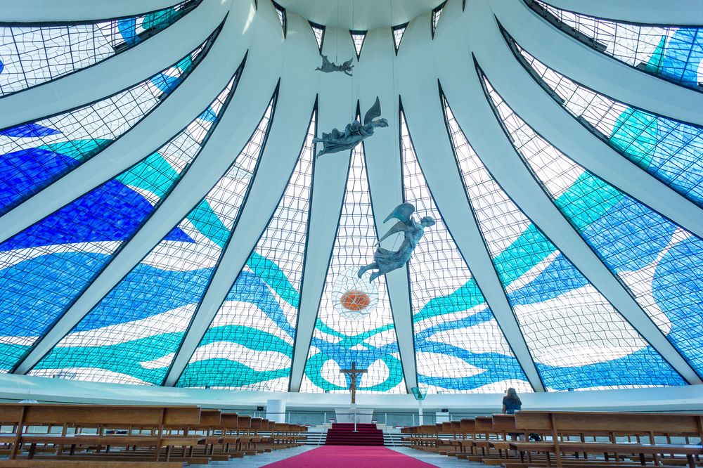 Modernist cathedral interior with dramatic blue and white stained glass windows in a geometric sunburst pattern.