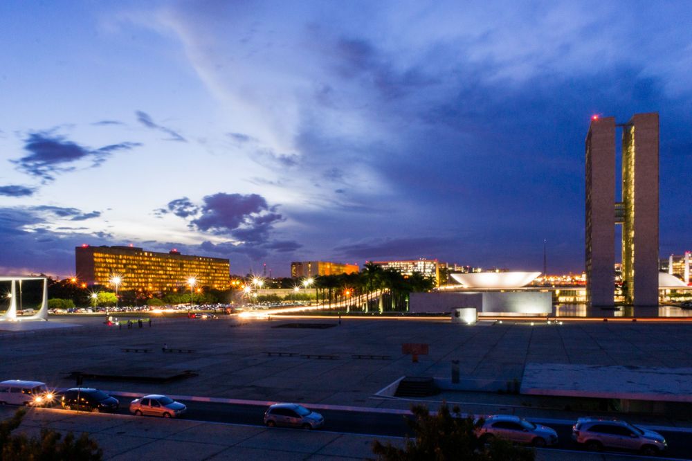 Panoramic twilight cityscape showing illuminated buildings along a waterfront with glowing lights reflecting on water.