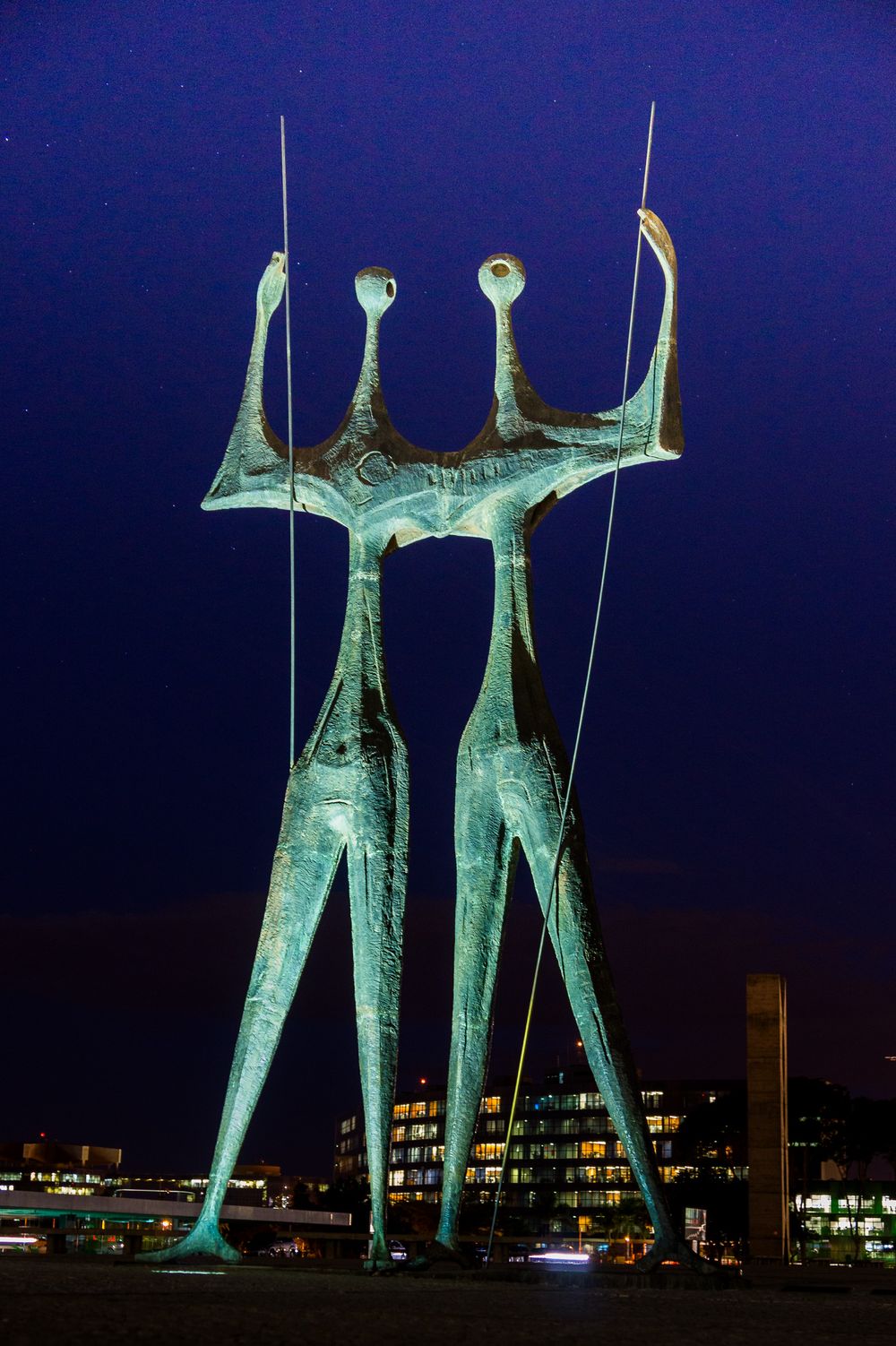 Illuminated green metal sculptures of figures standing together against a dark night sky in Brasilia's civic plaza.