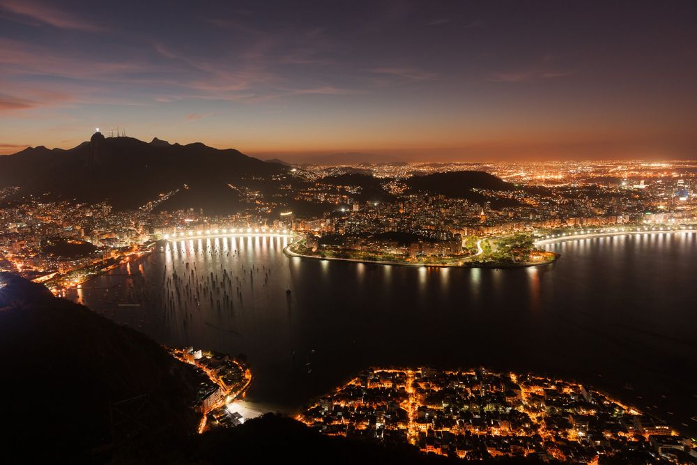 Aerial view of vibrant city lights reflecting in the bay at night with illuminated bridges and hills visible in the distance.