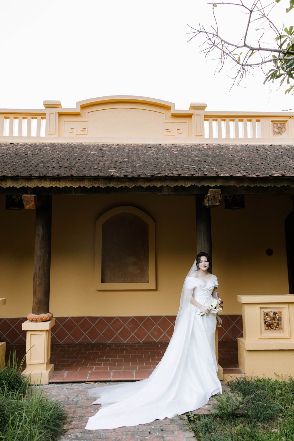 Bride in elegant white gown with long train stands before yellow colonial-style building exterior.