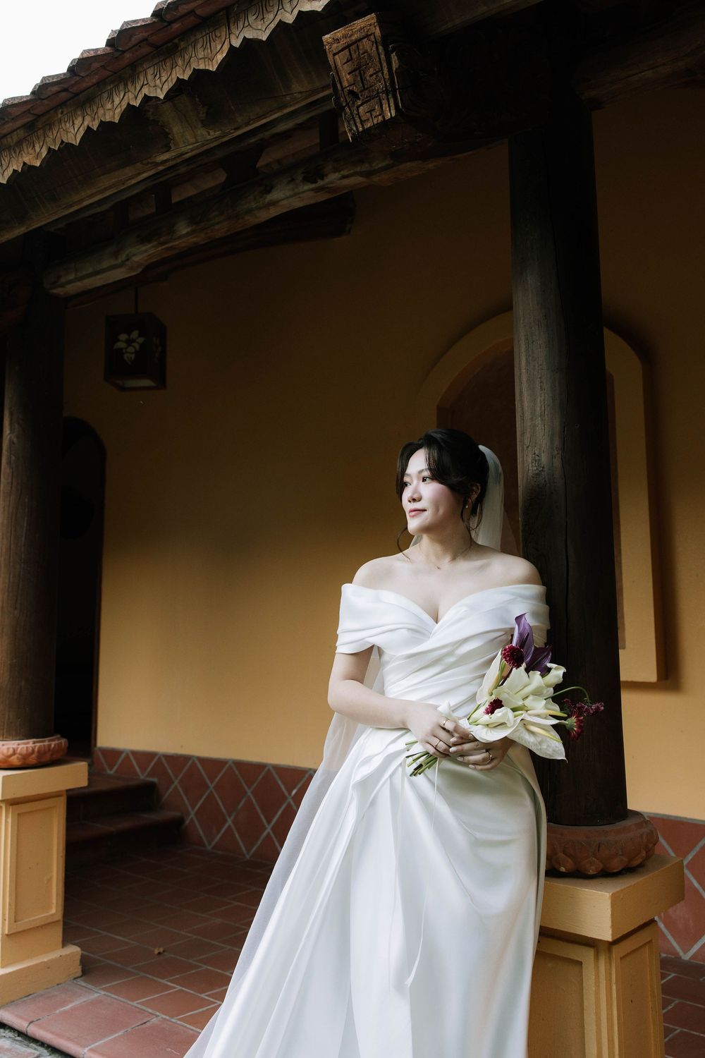 Bride in off-shoulder white gown holds floral bouquet beside ornate column of traditional Asian building.
