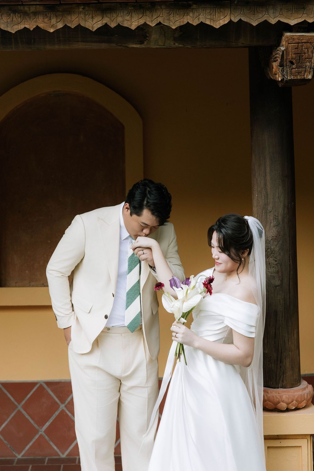 Groom in cream suit kisses bride's hand as she smiles holding bouquet by historic yellow building.