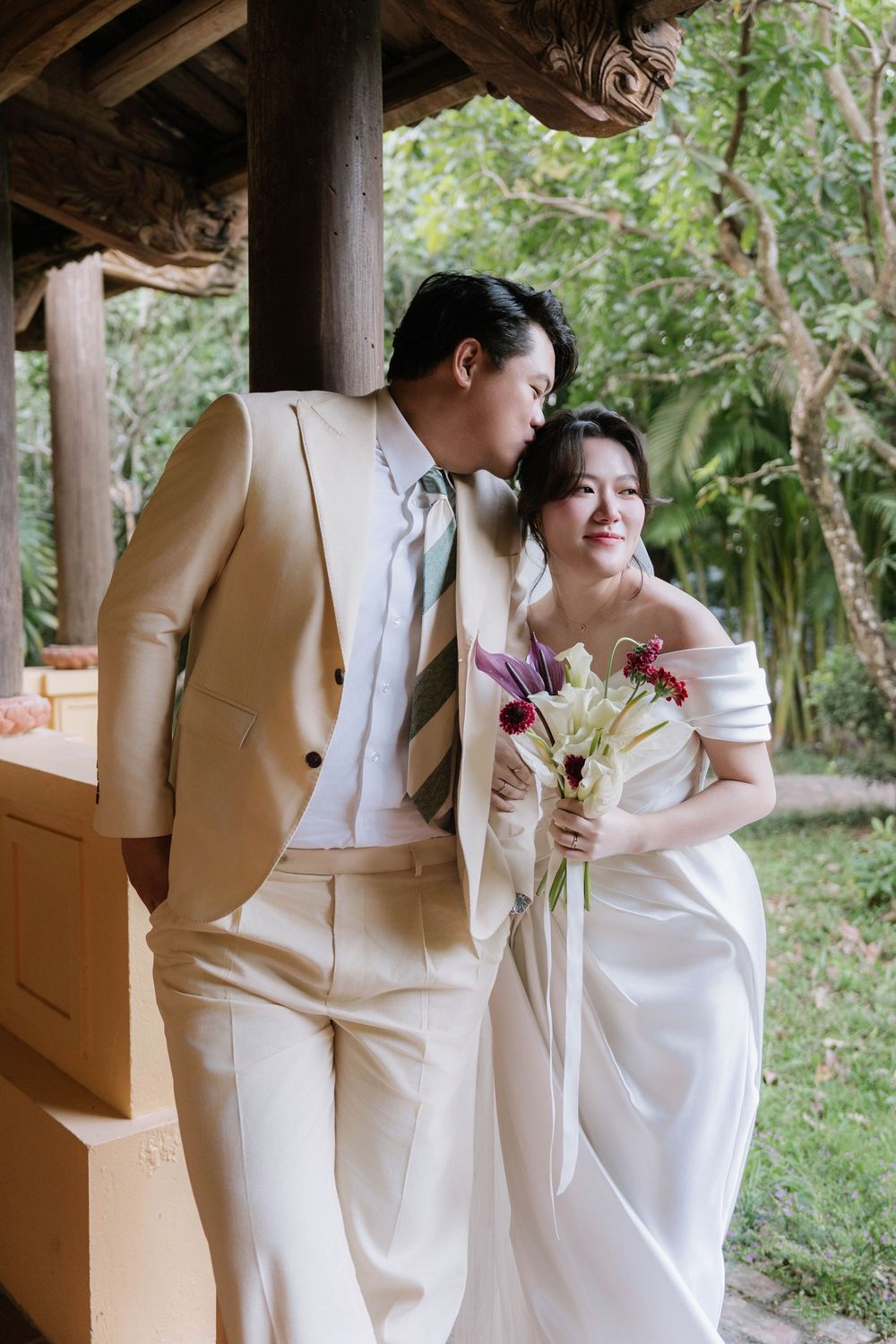 Groom in beige suit leans toward smiling bride holding colorful bouquet under garden pavilion.