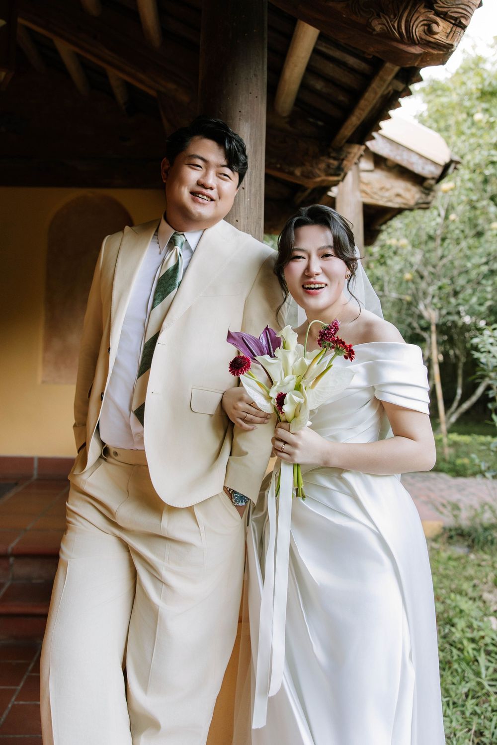 Smiling couple in wedding attire laugh together under wooden pavilion surrounded by lush greenery.