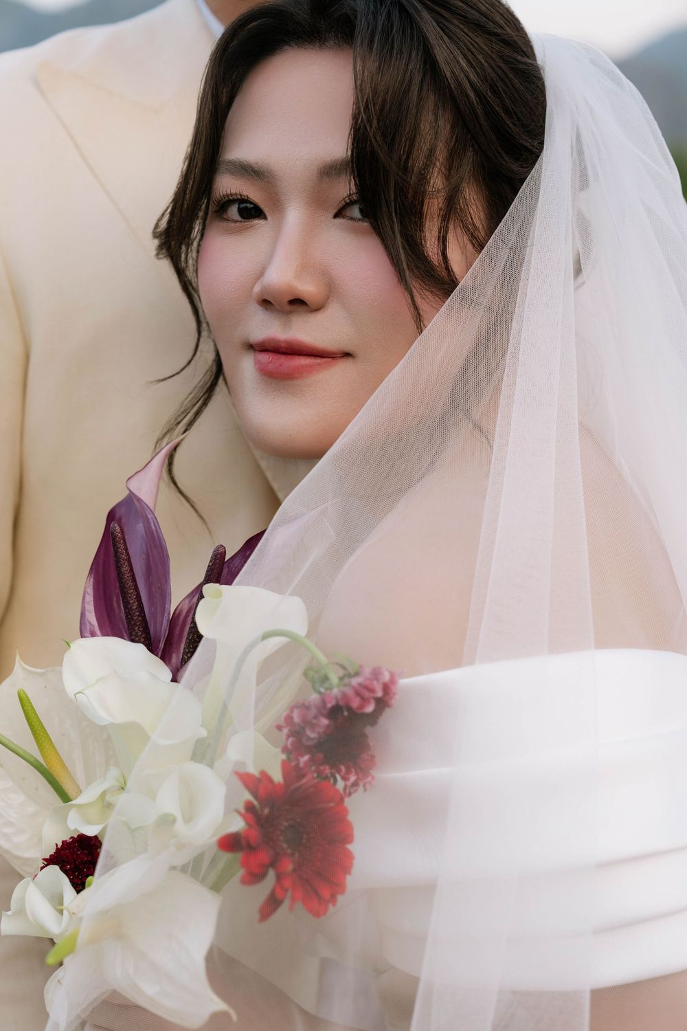Close-up portrait of smiling bride under veil holding white and red floral wedding bouquet.