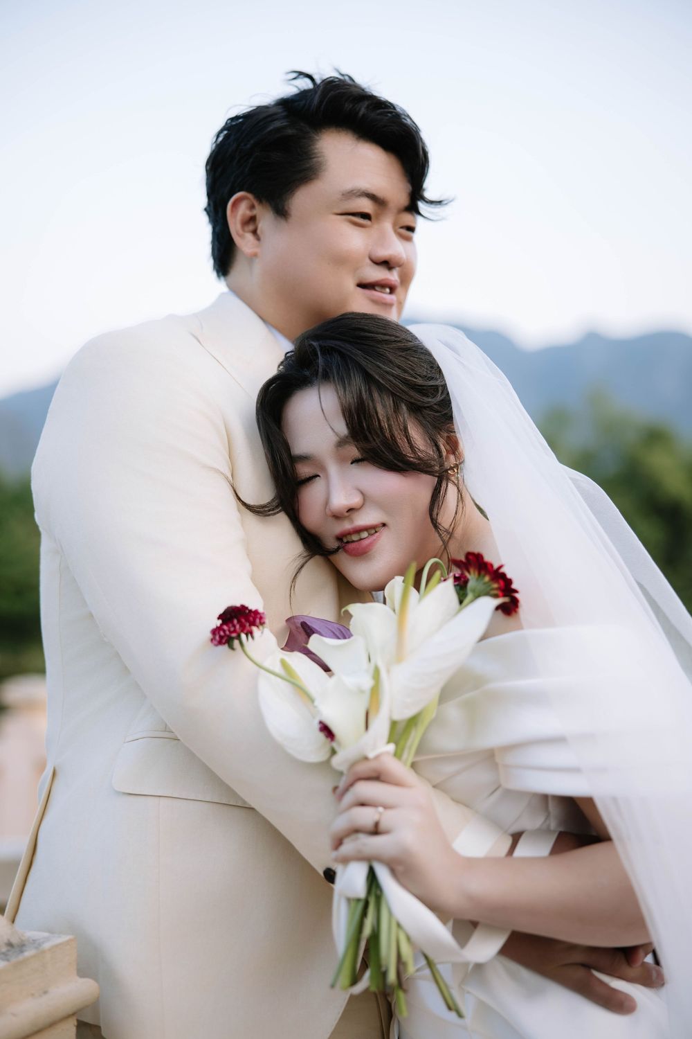 Groom embraces bride from behind as she smiles holding bouquet with scenic mountain backdrop.
