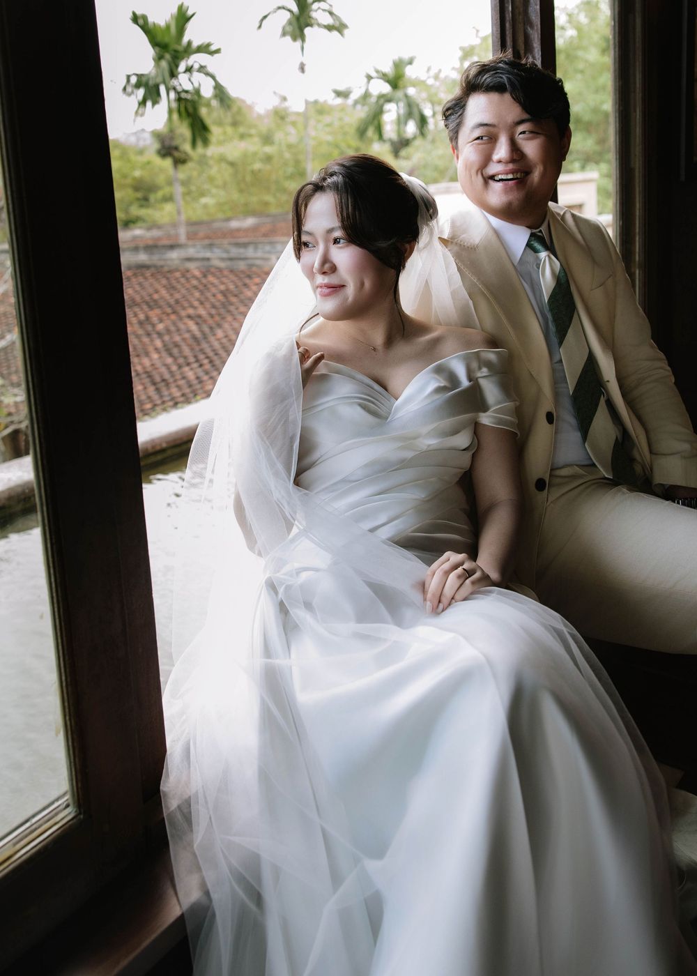 Bride and groom sit together by large window in vintage interior, smiling joyfully.