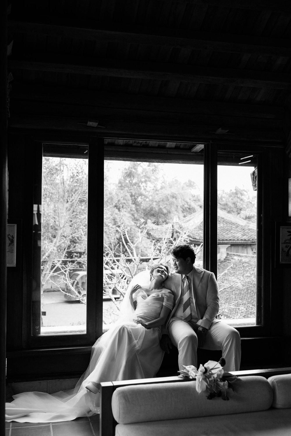 Black and white photo of laughing bride and groom seated by large window in dark interior.