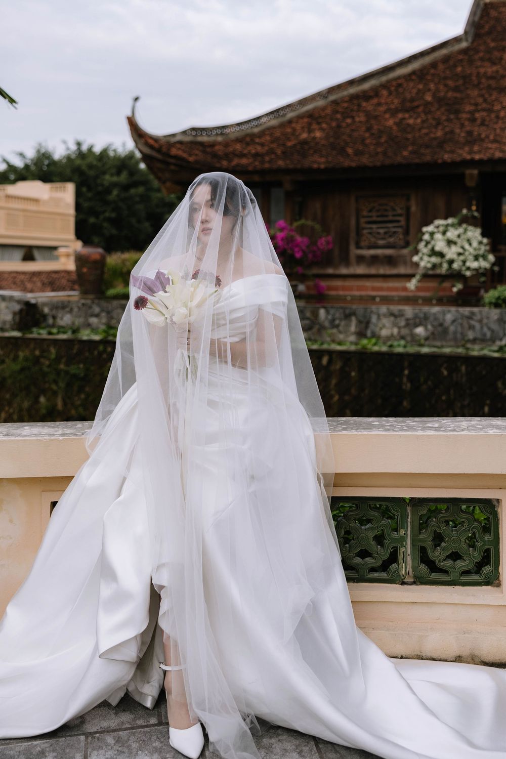 Bride in white off-shoulder gown and sheer veil holds bouquet at Asian temple.