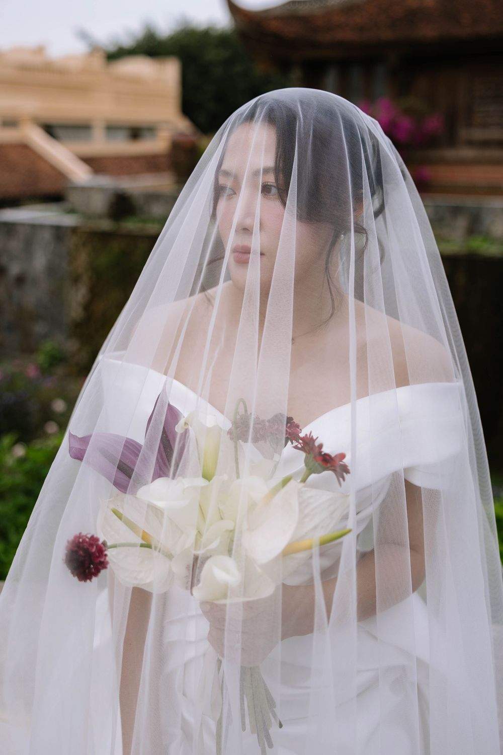 Beautiful bride under sheer veil holding white calla lily bouquet at outdoor venue.