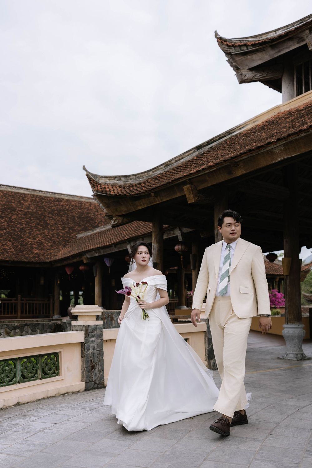 Couple in wedding attire walking hand in hand at traditional Asian architecture venue.