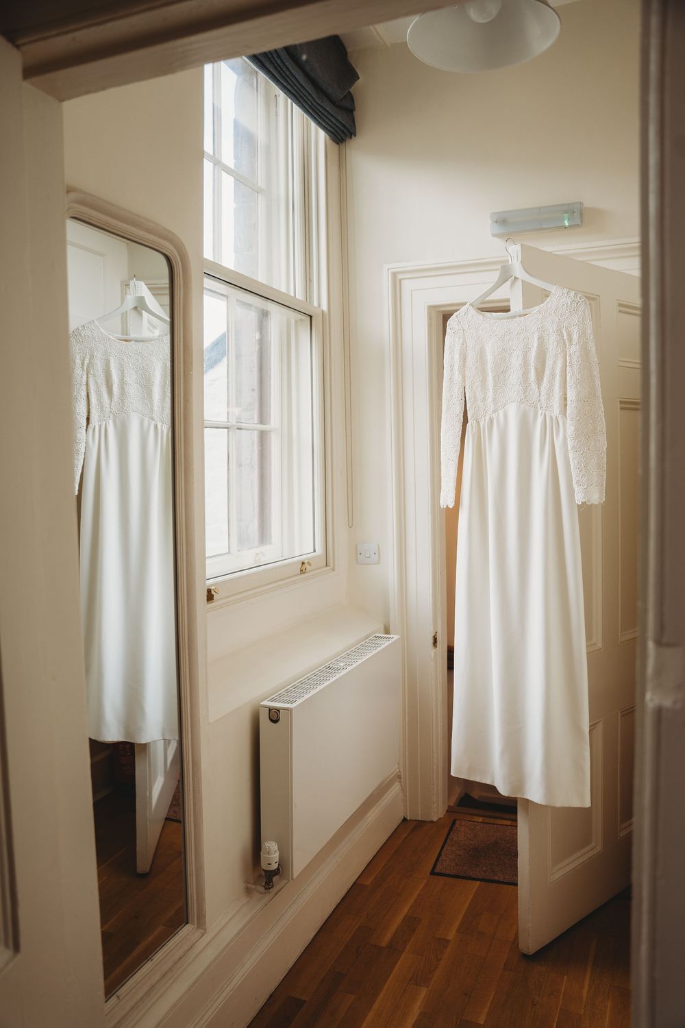 White wedding dress hanging by windows in a sunlit hallway with wooden floors.