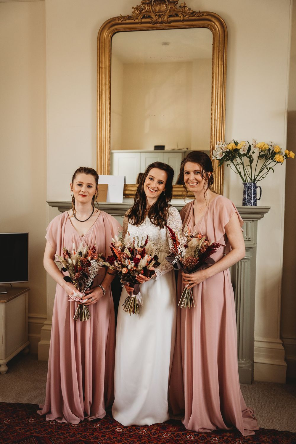 Wedding party in blush pink bridesmaid dresses and white bridal gown pose with bouquets in front of ornate gold mirror.