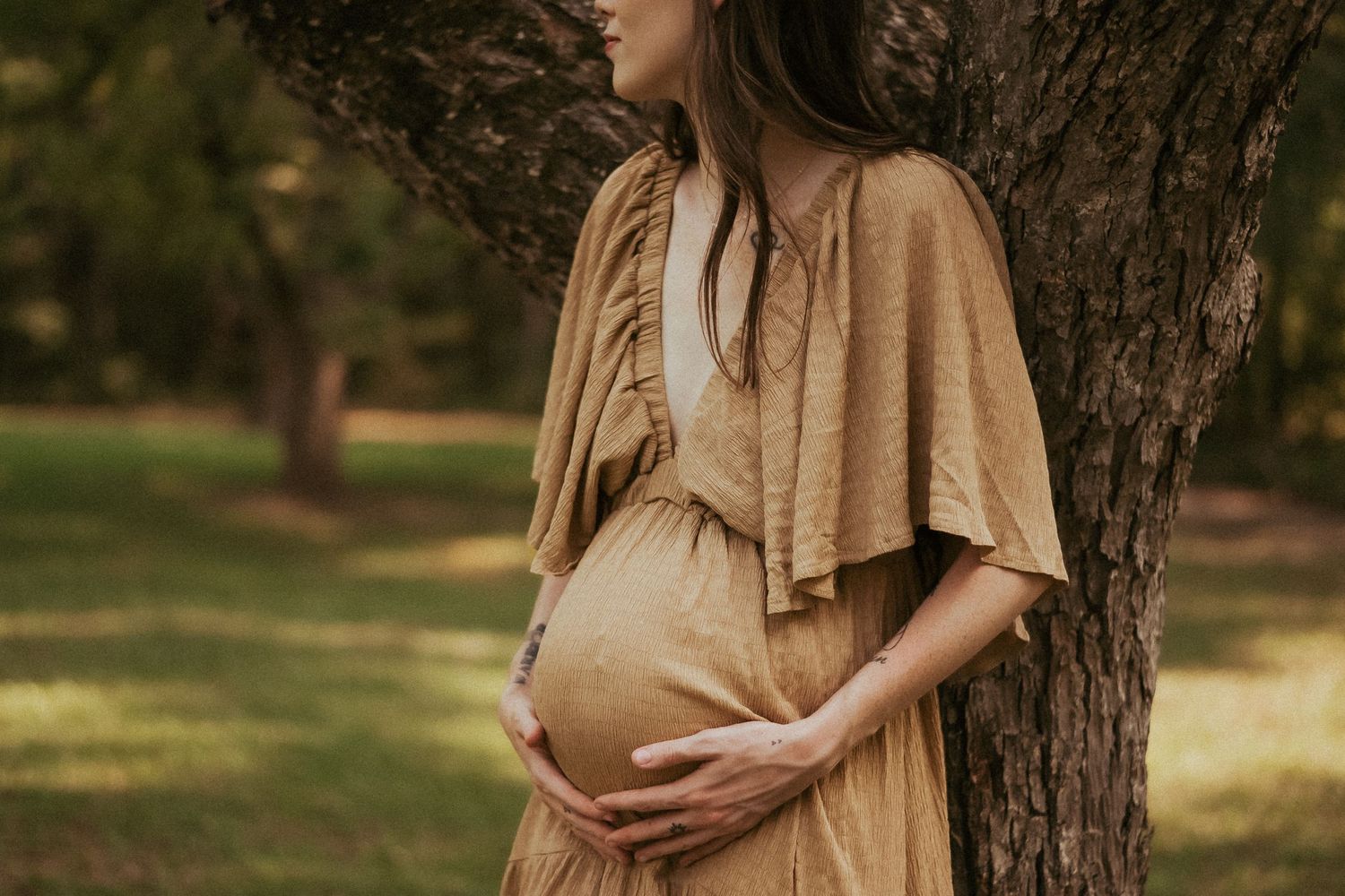 A woman in a flowing beige maternity dress stands against a tree while cradling her pregnant belly in a Rockingham, NC maternity session.