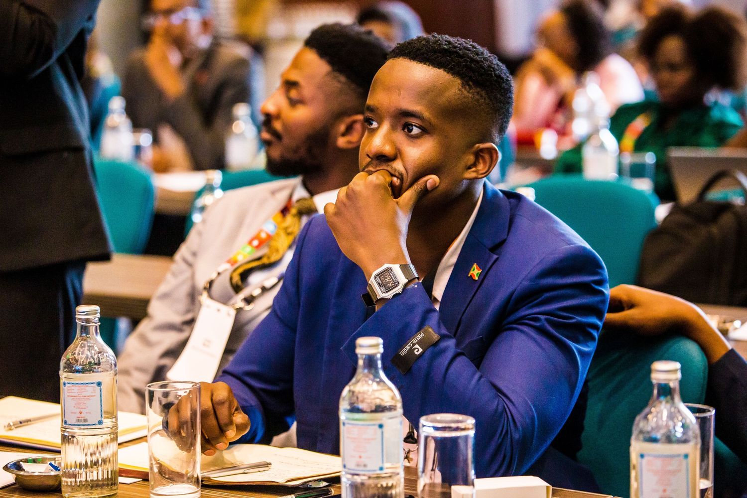Conference attendee in a navy blue blazer listens attentively while seated at a table with water bottles.