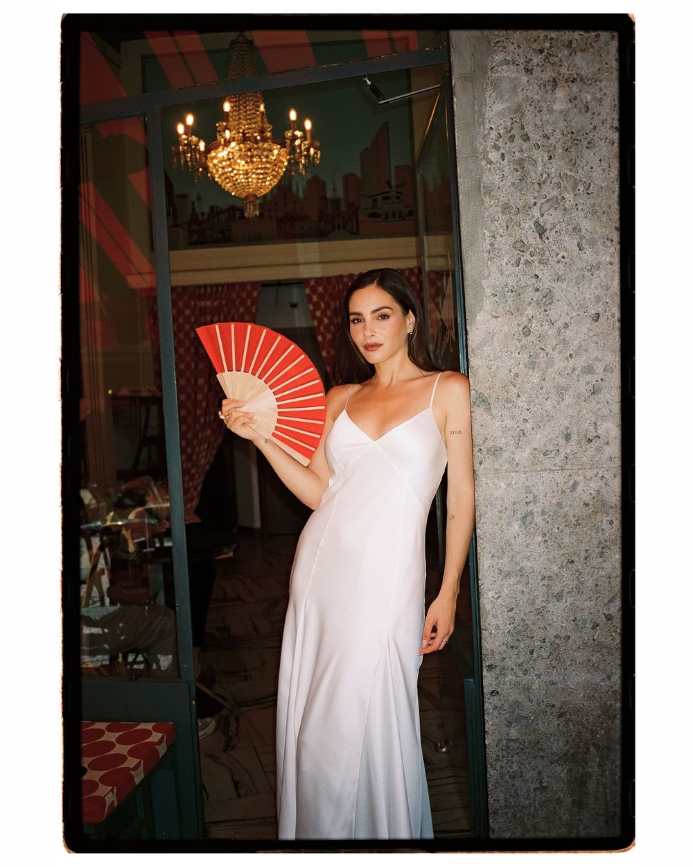 Woman in white slip dress holding red paper fan stands in doorway beneath crystal chandelier.