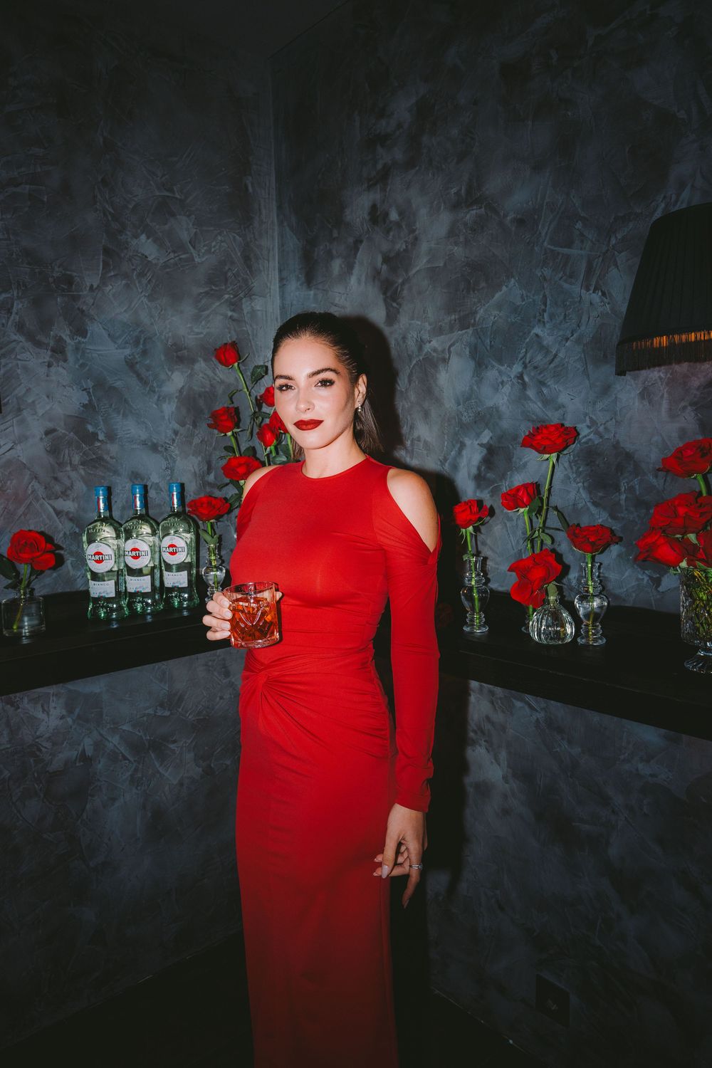 A figure in a striking red dress poses dramatically against a dark wall with red roses and glass bottles as decor.