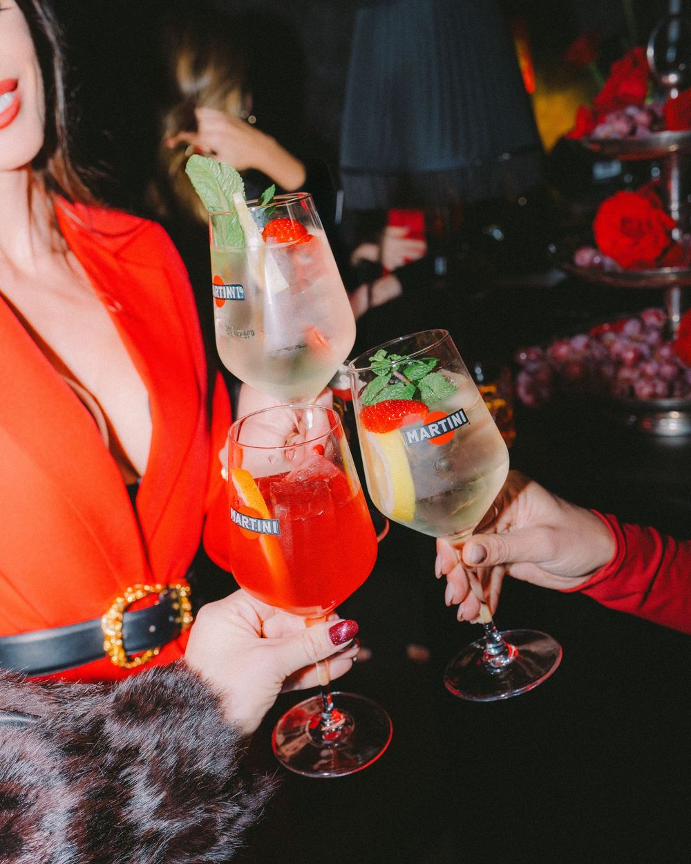 People holding cocktail glasses with beverages at a dimly lit bar or party setting.