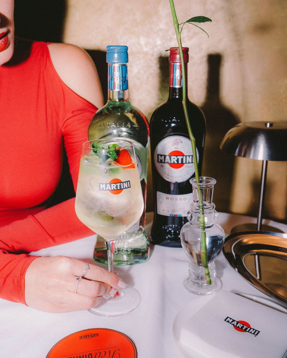 A person in a red top holds a glass of cocktail with garnish at a table with multiple liquor bottles in view.