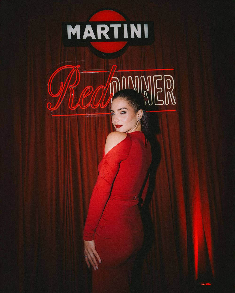 Figure in red dress poses dramatically against dark backdrop with illuminated Martini Red neon sign and red curtain.