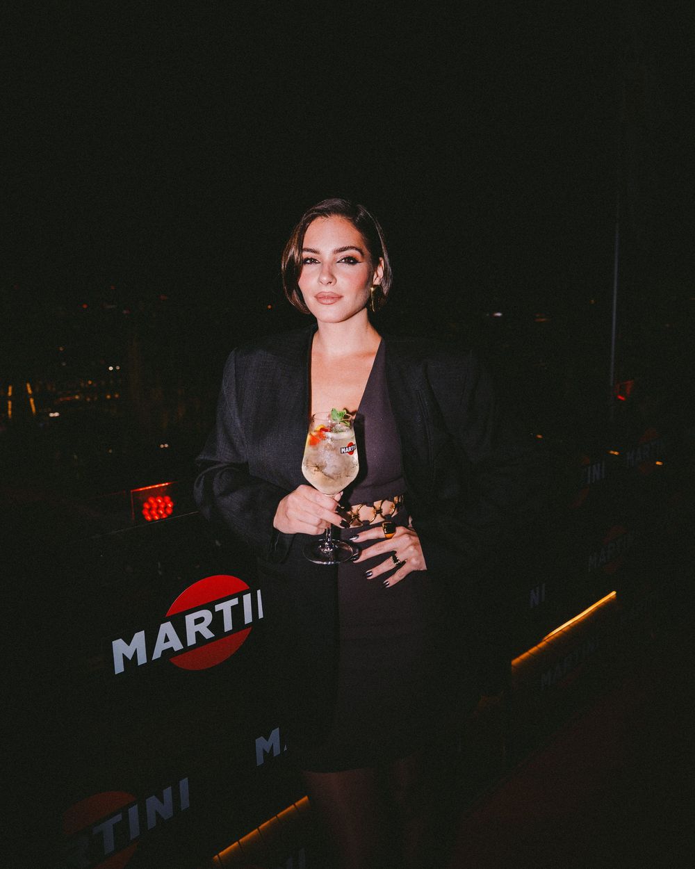 Person in dark setting holding a drink near illuminated Martini signage in moody nighttime atmosphere.