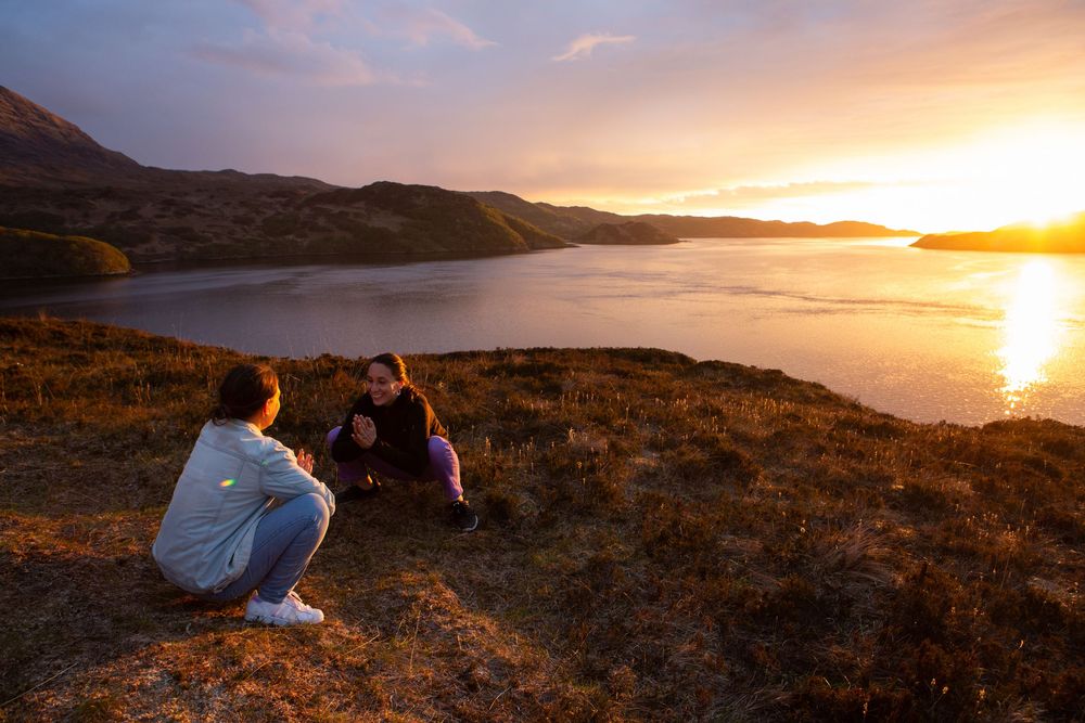 People camping and watching a stunning sunset over a scenic mountain lake with warm golden light reflecting on the water.
