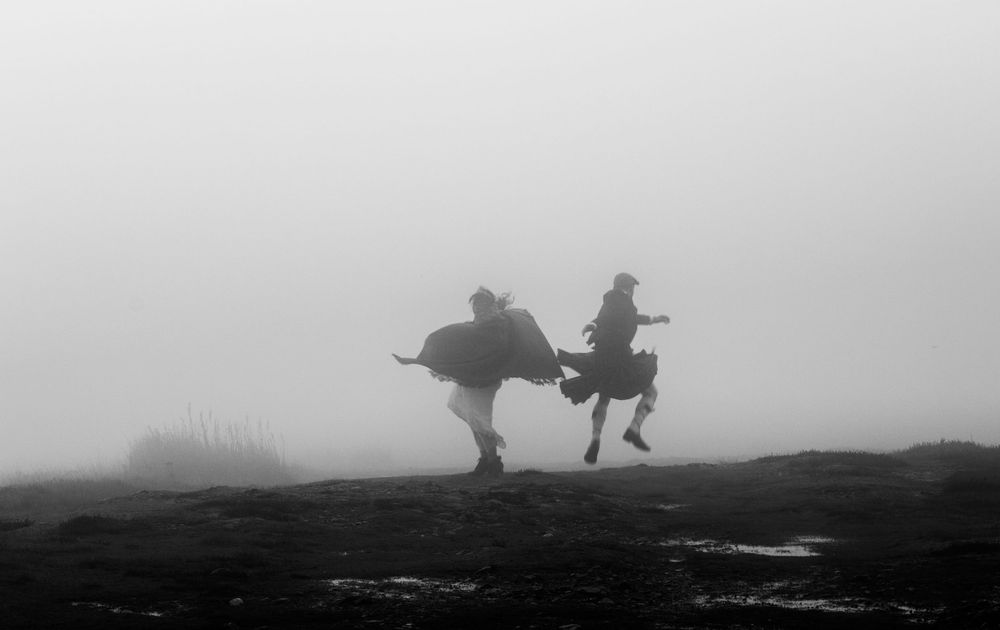 Two silhouetted figures leap joyfully through foggy terrain in a dramatic black and white photograph.