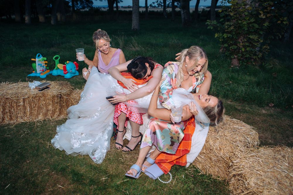 Wedding party members laughing together while sitting on hay bales at an outdoor rustic reception.