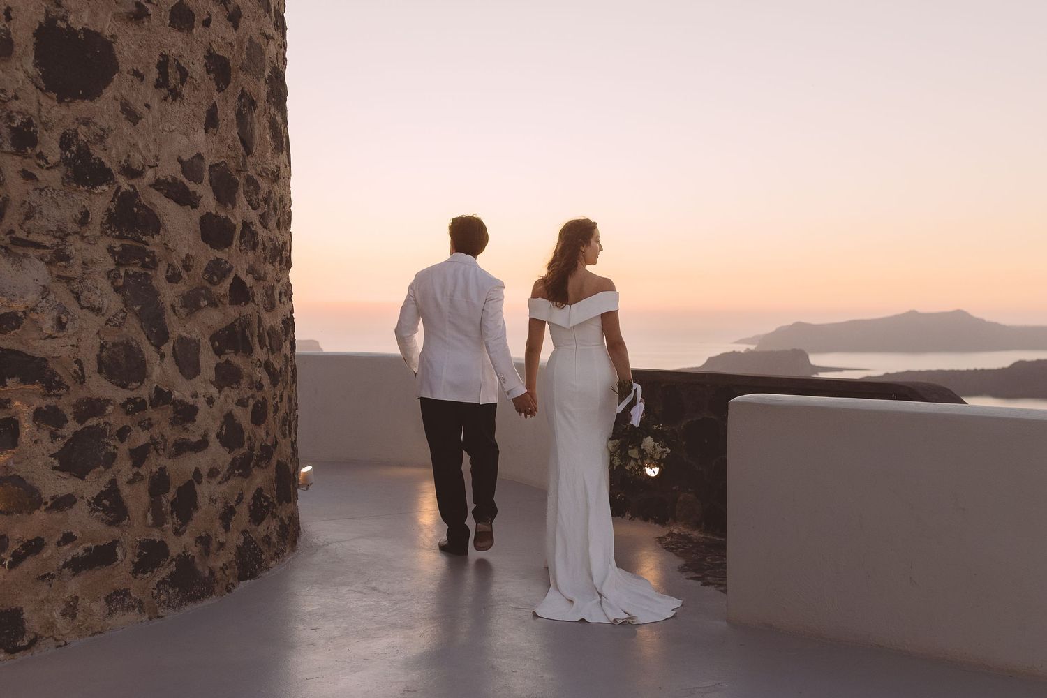 A couple in wedding attire stand on a stone terrace overlooking a sunset ocean view in Santorini, Greece.