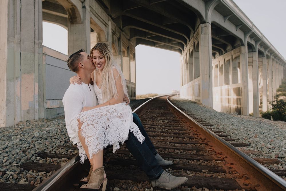 Couple laughing and embracing on railroad tracks under a bridge during a romantic outdoor engagement photo session.