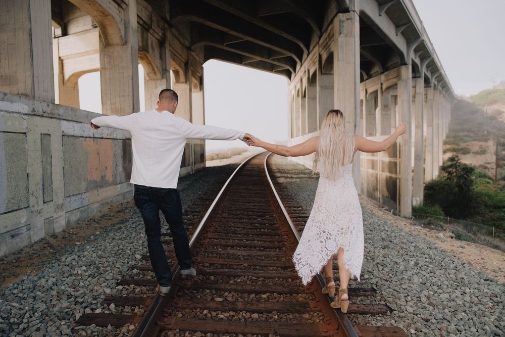 Couple holding hands walking on railroad tracks under a bridge, woman in white lace dress, man in dark pants.