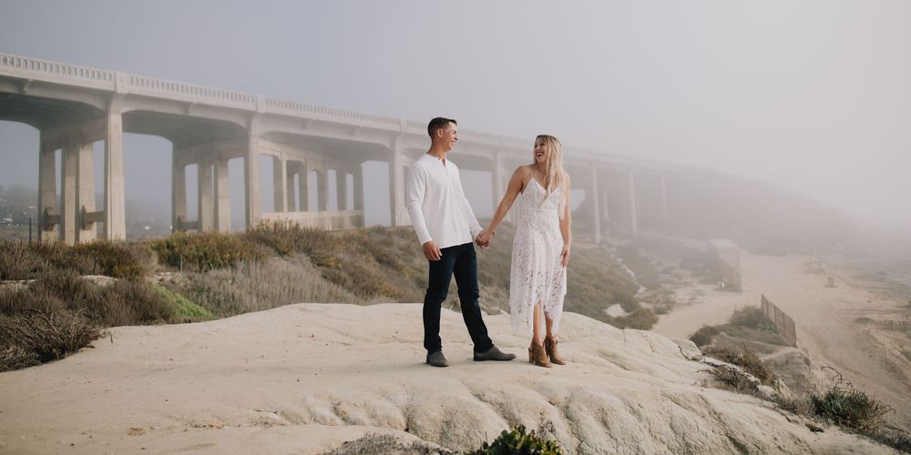 Couple holding hands on rocky coastal bluff with misty bridge backdrop during romantic outdoor engagement session.