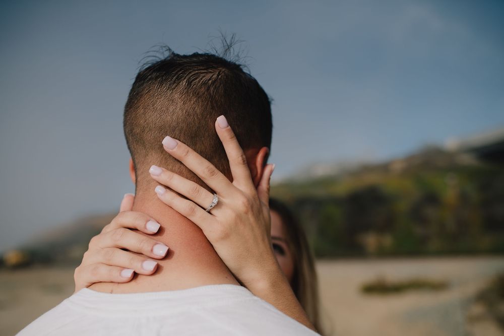 Woman with engagement ring embracing man outdoors on a sunny beach day.