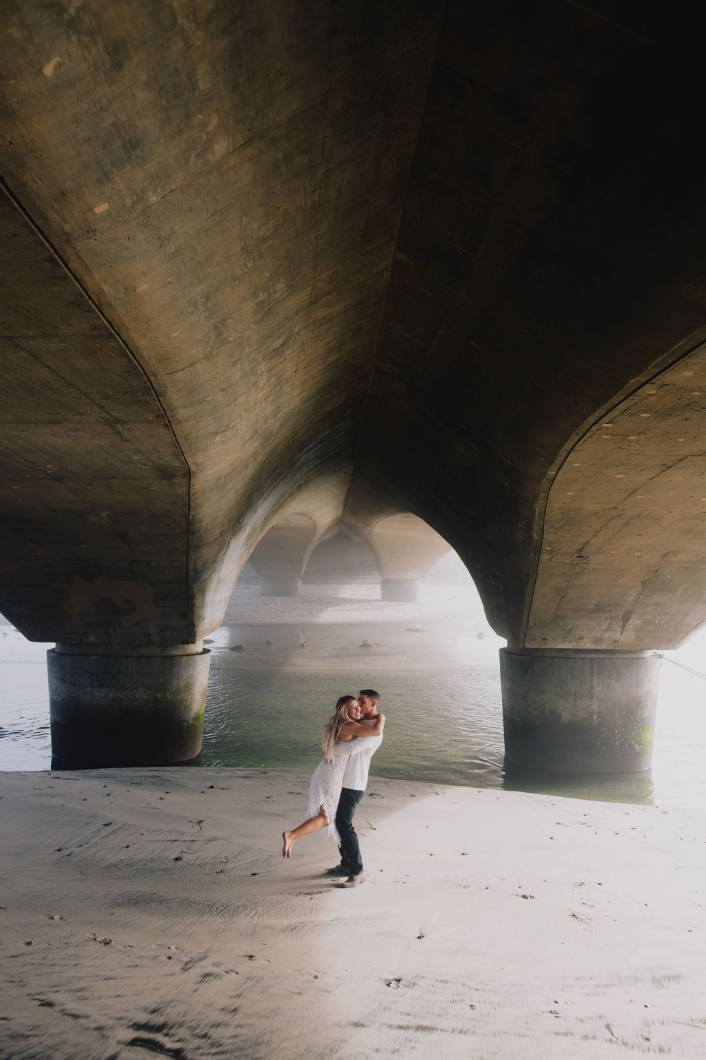 Couple embracing romantically under a stone bridge arch on sandy shore with water in the background.