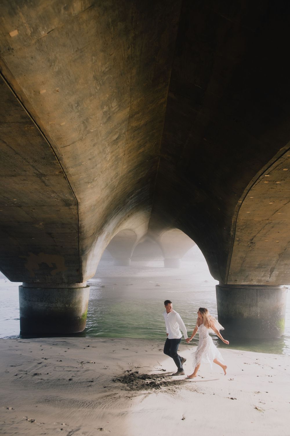 Couple running hand-in-hand on sandy beach beneath stone bridge arches with misty water in background.