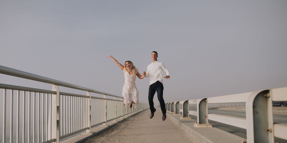 Joyful couple jumping and holding hands on a bridge pier on their wedding day with a clear sky backdrop.