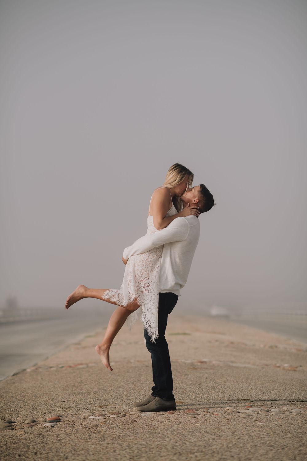 Man lifting and kissing woman in white lace dress on foggy beach road, romantic couple engagement photo.