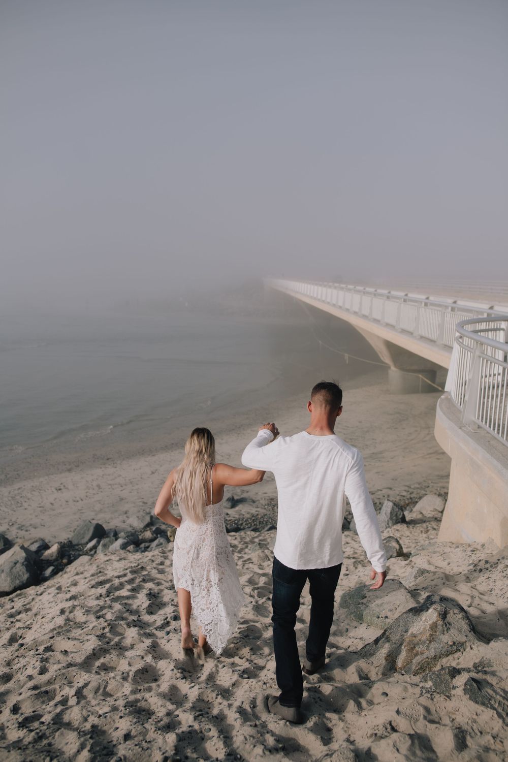 Couple walking hand in hand on a misty beach near a white bridge railing, woman in lace dress and man in white shirt.