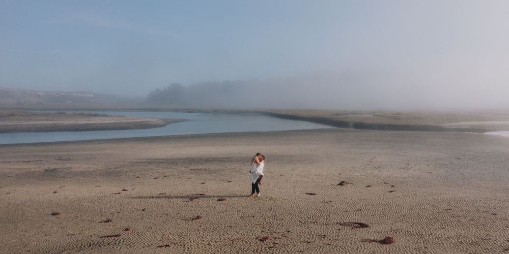 Person standing on a misty sandy beach with tidal flats and fog-covered hills in the background.