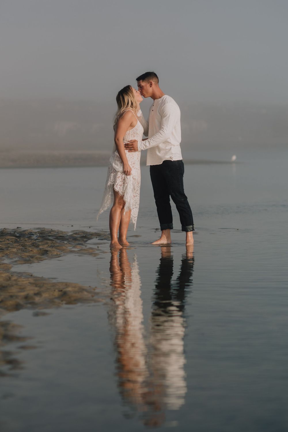 Couple sharing a romantic kiss while standing barefoot in shallow water on a misty beach.