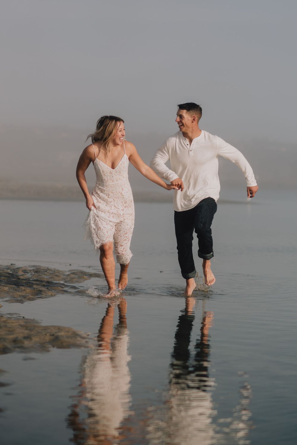 Couple in white outfits running barefoot through shallow beach water, laughing and holding hands on a misty day.
