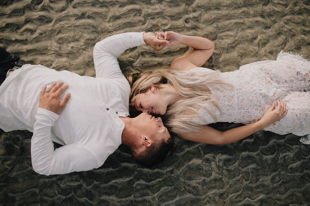 Romantic couple in white wedding attire lying on sandy beach, holding hands and gazing at each other.